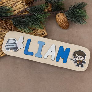 Könnte beinhalten: Ein Holzpuzzle mit dem Namen "LIAM" in blauen und weißen Buchstaben. Das Puzzle enthält ein Auto und eine Harry-Potter-Figur. Das Puzzle liegt auf einer hellbraunen Oberfläche mit Tannennadeln und Zapfen im Hintergrund.