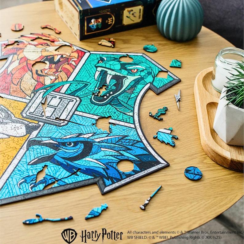 May include: A Harry Potter themed jigsaw puzzle featuring the Hogwarts crest with the four house mascots: a lion, a snake, an eagle, and a badger. The puzzle is partially completed and scattered pieces are visible on a wooden table.