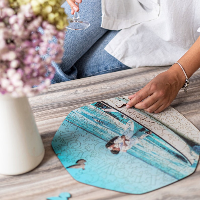 Peut inclure: Une personne assemble un puzzle en bois avec une photo d'un couple sur un bateau. Le puzzle est sur une table en bois. Les pi&egrave;ces du puzzle sont en forme de c&oelig;ur.
