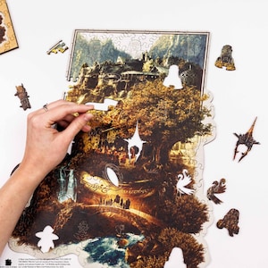May include: A partially assembled wooden jigsaw puzzle depicting a detailed landscape scene with a large tree and figures. Several puzzle pieces are scattered around, with a hand placing a piece. The puzzle has a fantasy theme.