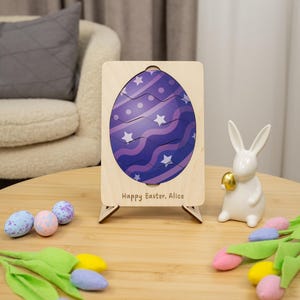 Peut inclure: Un puzzle en bois en forme d'œuf de Pâques avec un motif violet et blanc, posé sur une table en bois. Un lapin en céramique blanche tenant un œuf doré et des œufs de Pâques décoratifs sont à proximité. Le puzzle porte l'inscription "Happy Easter, Alice".
