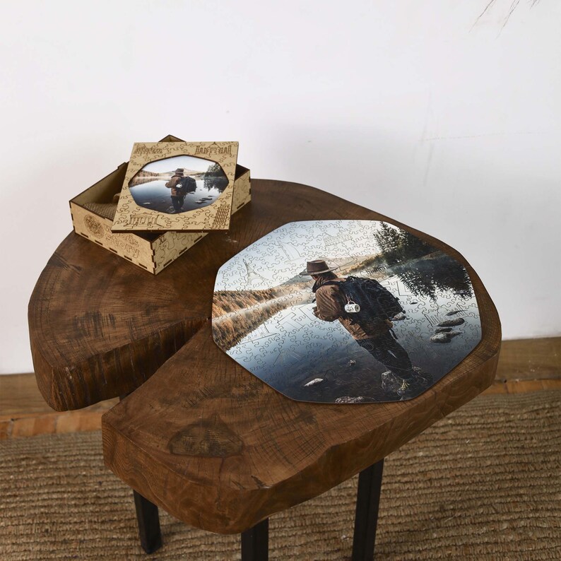 Op de afbeelding: Een houten legpuzzel met een foto van een persoon met een hoed die in een rivier staat. De puzzel is gedeeltelijk in elkaar gezet op een houten tafel. Een bijpassende houten doos met een ingelijste foto is ook aanwezig.