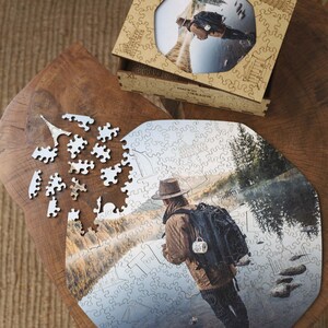 Op de afbeelding: Een houten legpuzzel met een foto van een persoon met een rugzak bij een water. De puzzel is gedeeltelijk in elkaar gezet op een houten oppervlak, met losse stukjes verspreid. Een houten doos met een bijpassende afbeelding is ook aanwezig. De tekst "MADE IN UKRAINE" is zichtbaar.