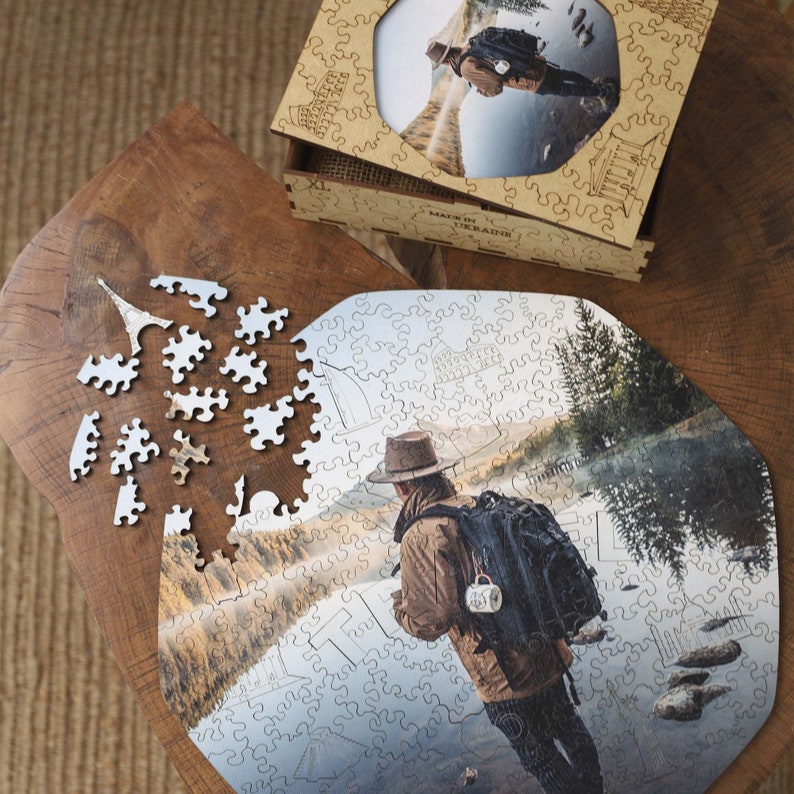 Op de afbeelding: Een houten legpuzzel met een foto van een persoon met een rugzak bij een water. De puzzel is gedeeltelijk in elkaar gezet op een houten oppervlak, met losse stukjes verspreid. Een houten doos met een bijpassende afbeelding is ook aanwezig. De tekst "MADE IN UKRAINE" is zichtbaar.