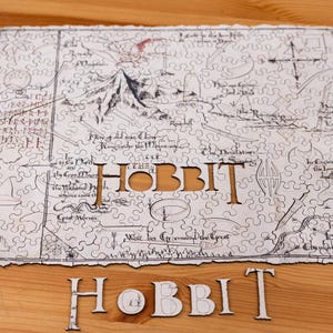 Puede incluir: Un rompecabezas de madera con un mapa con el texto "The Hobbit" recortado. Las piezas del rompecabezas son blancas con líneas negras, que representan un mapa detallado. La palabra "HOBBIT" también está escrita en letras de madera individuales.