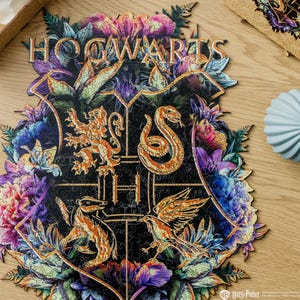 May include: A wooden jigsaw puzzle featuring the Hogwarts crest, surrounded by colorful floral designs. The crest includes a lion, snake, badger, and eagle, with the word "HOGWARTS" at the top. The puzzle is set against a wooden surface.