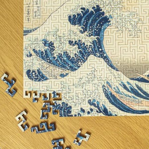 May include: A partially assembled jigsaw puzzle depicting Hokusai's "Great Wave off Kanagawa". The image shows a large blue wave with white foam, a mountain, and boats. Several puzzle pieces are scattered on a wooden surface, with the puzzle partially complete.