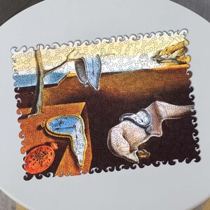 Op de afbeelding: Een houten legpuzzel met een surrealistisch schilderij met smeltende klokken, een boom en een landschap. De puzzel heeft een unieke, golvende rand en wordt op een witte tafel getoond.