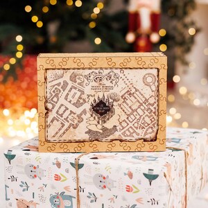 May include: A wooden puzzle box featuring a map design with the text "The Marauders Map". The box is sitting on top of a wrapped gift with a colorful pattern. The background is blurred with Christmas lights.