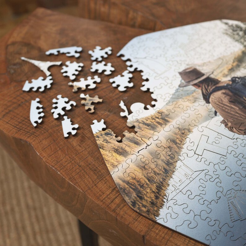 Op de afbeelding: Een gedeeltelijk voltooid legpuzzel met een landschapsafbeelding, met een persoon met hoed en rugzak. Verschillende puzzelstukjes liggen verspreid op een houten oppervlak. De puzzelstukjes zijn wit en bruin.