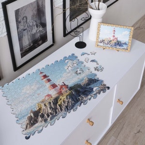May include: A completed jigsaw puzzle of a red and white lighthouse on a rocky cliff with blue water and a cloudy sky. The puzzle is laid out on a white table with a few pieces missing.