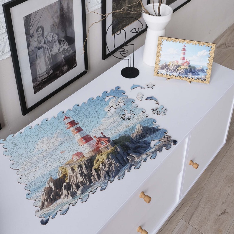 May include: A completed jigsaw puzzle of a red and white lighthouse on a rocky cliff with blue water and a cloudy sky. The puzzle is laid out on a white table with a few pieces missing.