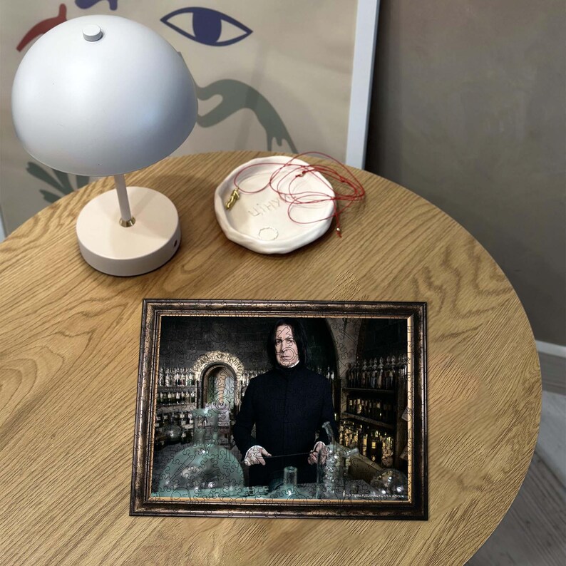 Puede incluir: Una imagen enmarcada de un hombre con el pelo largo y negro y una t&uacute;nica negra de pie en una habitaci&oacute;n con muchas botellas. El hombre sostiene una varita en su mano derecha. La imagen est&aacute; sobre una mesa de madera con una l&aacute;mpara blanca y un plato blanco con una cuerda roja.
