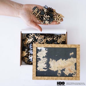 May include: A wooden jigsaw puzzle featuring a map design, with pieces held above the open box. The puzzle pieces are in shades of brown and black. The box has a map image on the bottom. The HBO logo is visible.