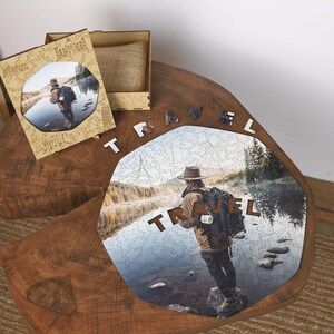 Op de afbeelding: Een houten legpuzzel met een foto van een persoon met een rugzak bij een meer. Het woord "TRAVEL" is in houten letters gespeld. Een houten doos en een ingelijste foto zijn ook aanwezig.
