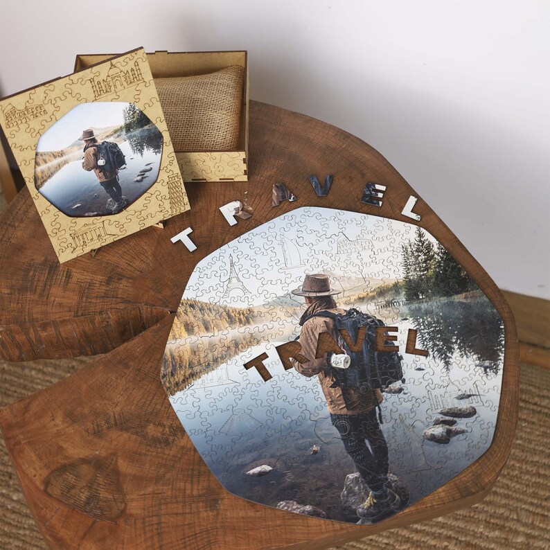 Op de afbeelding: Een houten legpuzzel met een foto van een persoon met een rugzak bij een meer. Het woord "TRAVEL" is in houten letters gespeld. Een houten doos en een ingelijste foto zijn ook aanwezig.