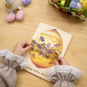 May include: A wooden Easter egg puzzle with a yellow and orange design, featuring purple floral accents. The puzzle is being assembled by a child. The text "Happy Easter, Alice" is printed on the puzzle. Decorative Easter eggs and a basket are in the background.