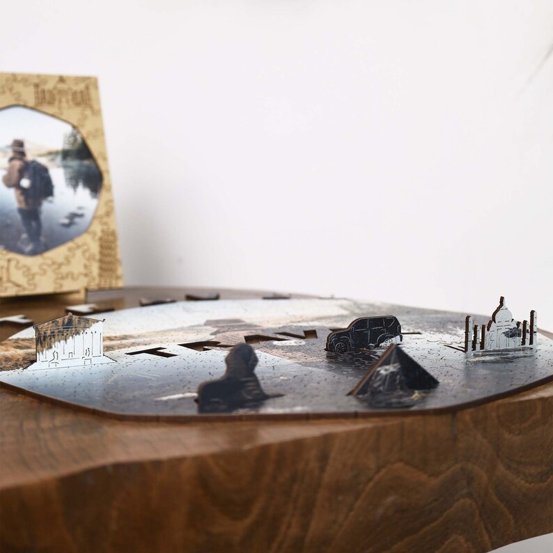 Op de afbeelding: Een houten legpuzzel met lasergesneden stukken, met miniatuur bezienswaardigheden zoals de Sfinx, een auto en een tempel. De puzzel is gedeeltelijk gemonteerd op een houten oppervlak, met een ingelijste foto op de achtergrond.