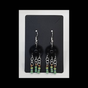 May include: A pair of unique earrings featuring black gear-shaped pendants. Each earring has multiple dangling chains with small, colorful cylindrical beads. The earrings are displayed on a black card with a white background.