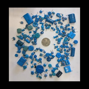 May include: A collection of various blue and teal electronic capacitors of different shapes and sizes, scattered on a white surface. A quarter coin is in the center for size reference. The capacitors have metal leads.