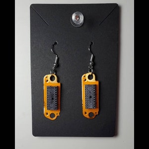 May include: A pair of unique dangle earrings. Each earring features a rectangular, orange-colored circuit board with a black integrated circuit chip. The earrings hang from silver-colored fish hook ear wires, displayed on a black card.