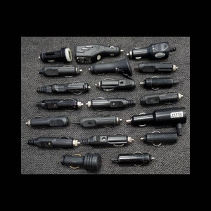 May include: A collection of black 12-volt DC car charger adapters, each with a different design. The adapters are made of plastic and metal, and are arranged on a gray textured surface. Some have indicator lights.