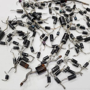 May include: A collection of black electronic diodes with silver leads scattered across a white surface. The diodes have various markings and are cylindrical with a black body and silver leads. Some diodes have a brown or orange band.