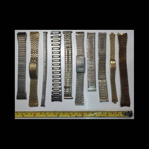 May include: A collection of vintage watch bands in various metallic tones, including silver and gold. The bands are of different styles and widths, with a measuring tape at the bottom.