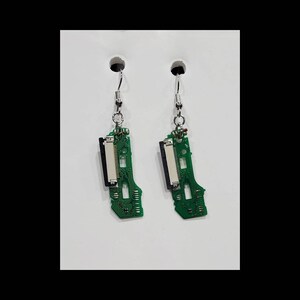 May include: A pair of dangle earrings featuring repurposed green circuit board fragments as the main pendants. Each piece includes a silver-toned metal connector and a standard silver-toned hook wire for attachment, showcasing intricate electronic traces.