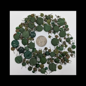 May include: A collection of green and olive-colored electronic components, including disc capacitors, arranged in a circular pattern. A U.S. nickel coin is in the center for size comparison. The components have wire leads and various markings.