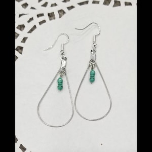 May include: A pair of silver teardrop earrings with a small turquoise bead accent. The earrings have a simple, elegant design with a hook closure. The earrings are displayed on a white, textured surface.