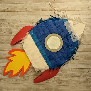 May include: A blue, white, and red rocket-shaped pinata with a silver circle in the center. The pinata is hanging from a string and has a yellow flame coming out of the back.
