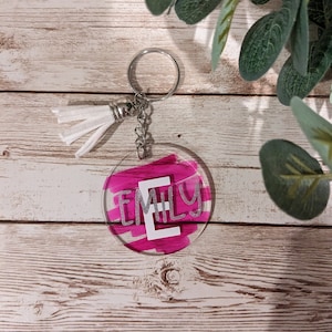May include: A pink and white round keychain with the name "Emily" written in silver glitter. The keychain has a white tassel attached to a silver key ring.