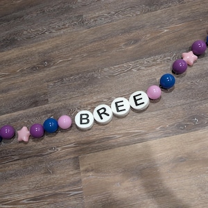 May include: A decorative arrangement on a wooden surface. The word "BREE" is spelled out with white circular tiles with black letters. The letters are interspersed with pink, purple, and blue spheres and a pink star-shaped bead.