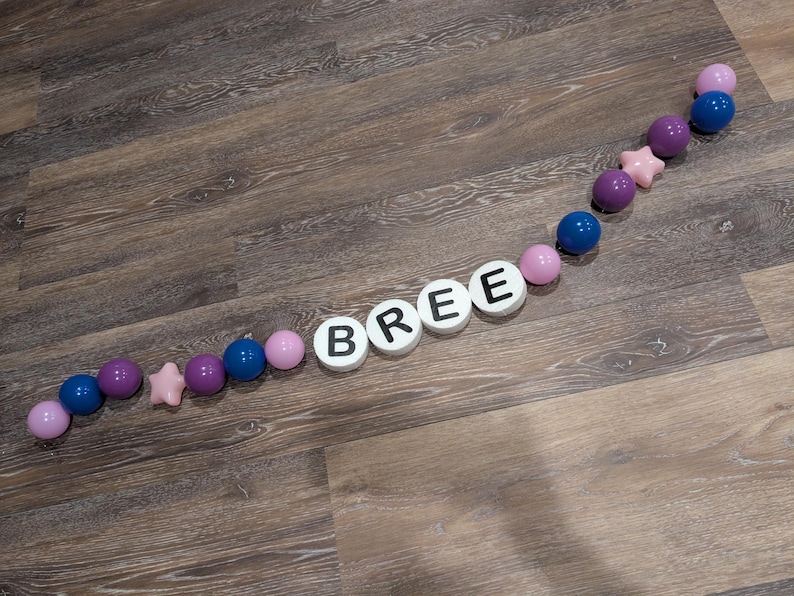 May include: A decorative arrangement on a wooden surface. The word "BREE" is spelled out with white circular tiles with black letters. The letters are interspersed with pink, purple, and blue spheres and a pink star-shaped bead.
