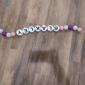 May include: A decorative garland spelling out the name "BLAKLEY" with white letter tiles and colorful beads. The beads are pink, white, and purple. The garland is arranged on a wooden surface.