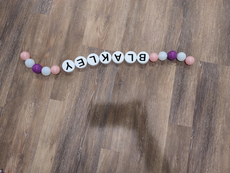 May include: A decorative garland spelling out the name "BLAKLEY" with white letter tiles and colorful beads. The beads are pink, white, and purple. The garland is arranged on a wooden surface.