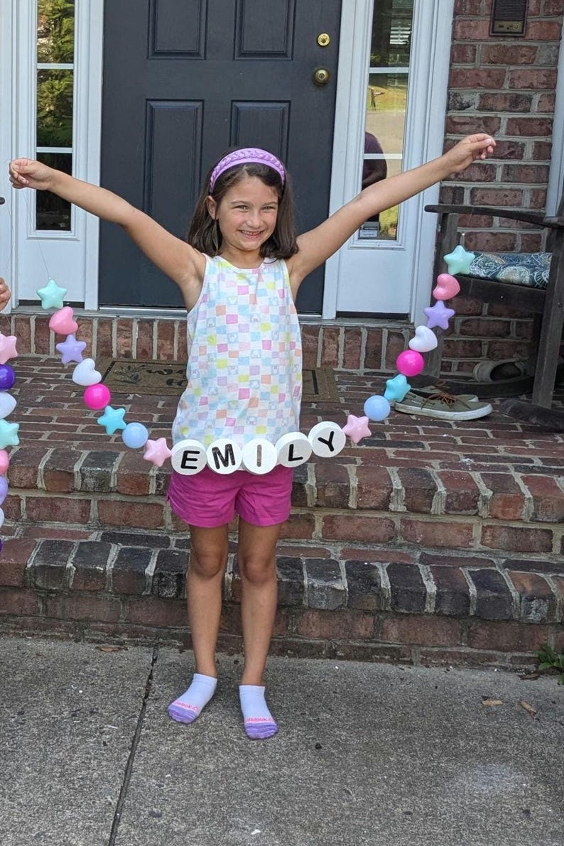 May include: A young person wearing a white tank top with a colorful checkered pattern and pink shorts. A beaded garland spelling "EMILY" is held up. The garland includes star and heart-shaped beads in pastel colors.