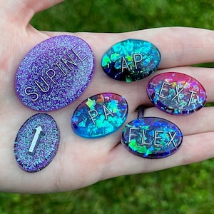May include: A set of six iridescent, glittery, oval-shaped resin tokens with various words and numbers printed on them. The words include "SUPINE", "AP", "EXT", "PA", "FLEX", and the number "1".