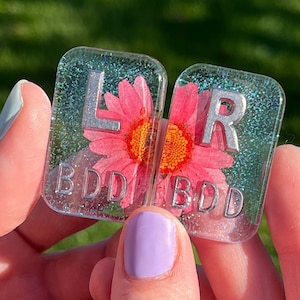 May include: Two clear resin dice with silver letters and a pink flower pressed inside. The dice read "L BDD" and "R BDD".