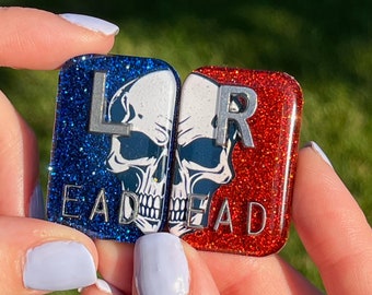 May include: Two square-shaped, glitter-covered, skull-shaped charms with the letters "L" and "R" on them. The left charm is blue with the word "DEAD" underneath the skull. The right charm is red with the word "DEAD" underneath the skull.