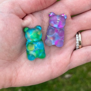 May include: Two colorful gummy bear-shaped charms. One is blue and green, the other is purple and pink. The charms are translucent and have a glossy finish. They are held in a hand, with a blurred green background.