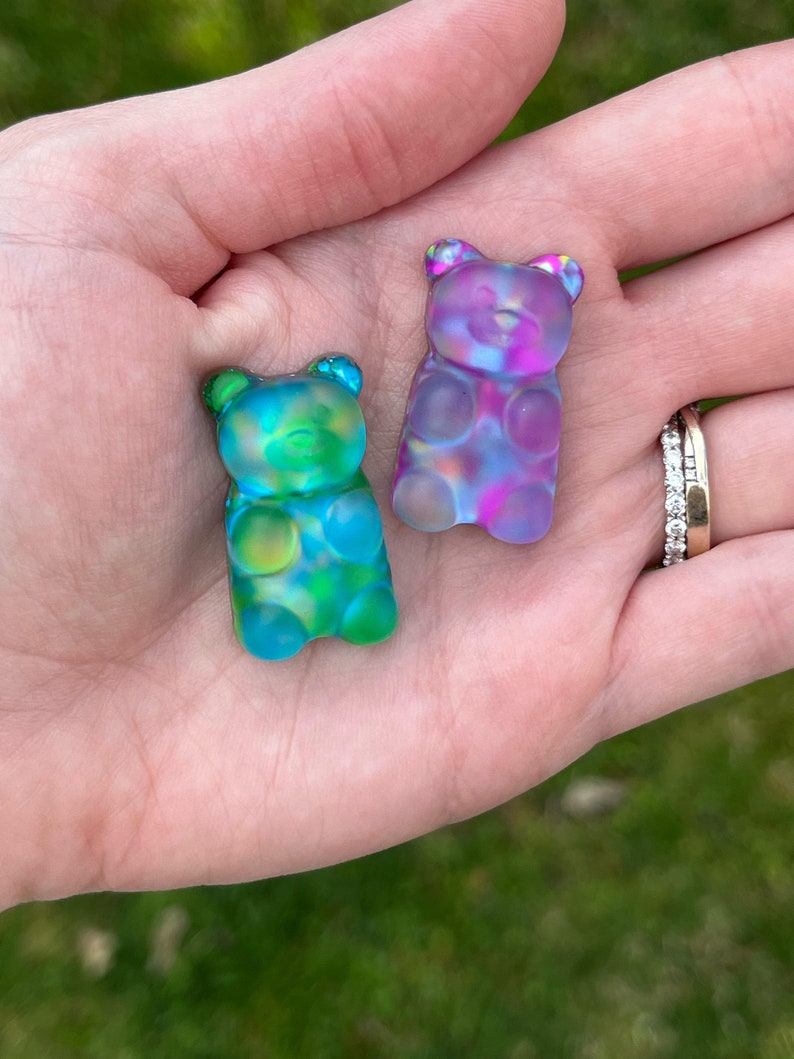 May include: Two colorful gummy bear-shaped charms. One is blue and green, the other is purple and pink. The charms are translucent and have a glossy finish. They are held in a hand, with a blurred green background.