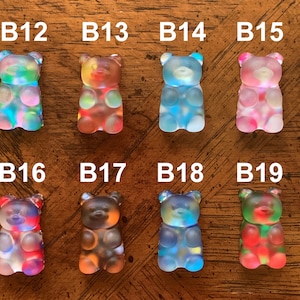 May include: A collection of eight colorful gummy bear figurines. Each bear is a different color, including blue, pink, red, and green. The bears are labeled with the letters "B" followed by a number.