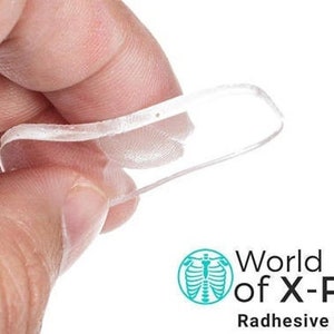 May include: A clear, flexible Radhesive Strip is held between fingers. The product is from World of X-Ray. The strip is thin and appears to be made of a transparent material. The image is set against a white background.