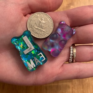 May include: Two resin gummy bear charms. One is blue with silver glitter and the letters "MAE". The other is a translucent purple and pink. A quarter coin is also in the image.