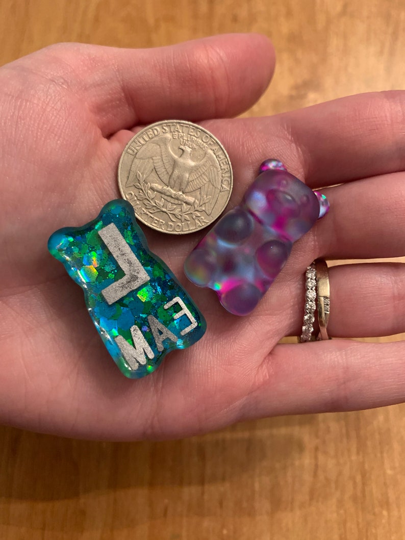 May include: Two resin gummy bear charms. One is blue with silver glitter and the letters "MAE". The other is a translucent purple and pink. A quarter coin is also in the image.