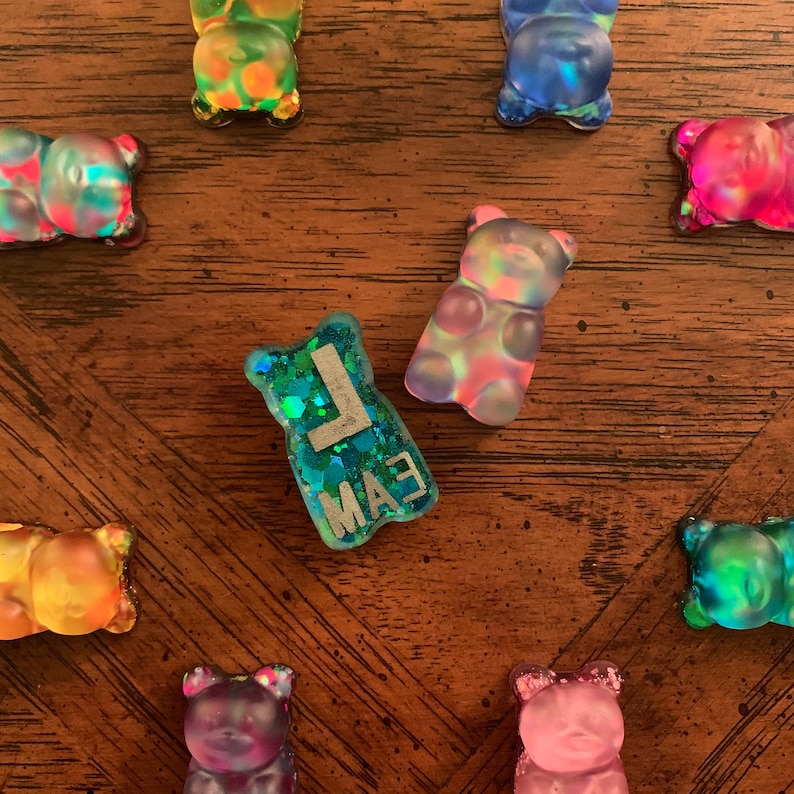 May include: A collection of colorful, translucent gummy bear-shaped resin charms. One charm is teal with silver glitter and the letters "LMA3". The other charms are various colors, including pink, blue, green, and yellow, with iridescent effects.