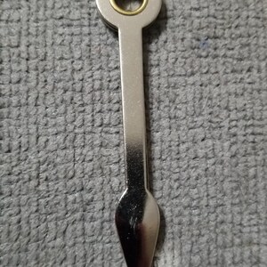 May include: A silver-colored metal wine bottle opener with a circular top featuring a gold-colored ring. The opener has a long, slender body that tapers to a pointed end with a small, circular base.