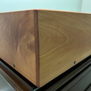 May include: Close-up of a wooden box with a natural wood finish. The box has a smooth surface and is made of solid wood. The box is sitting on a dark brown surface.